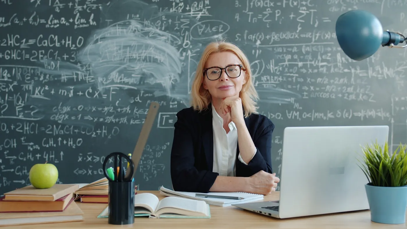 A teacher at her desk in front of a chalkboard, smiling with her chin resting on her hand